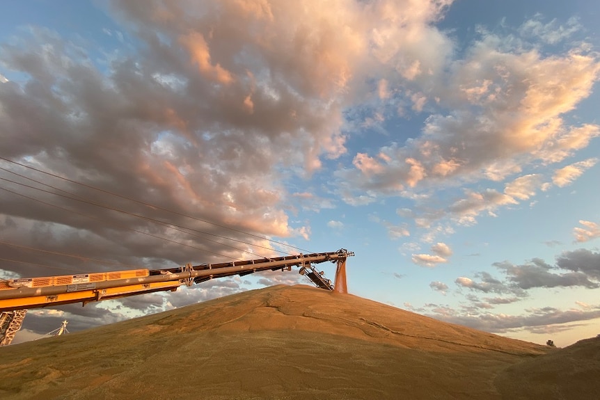 The sun is setting creating colourful skies above a large pile of grain. A long mechanical arm spurting more into the pile.