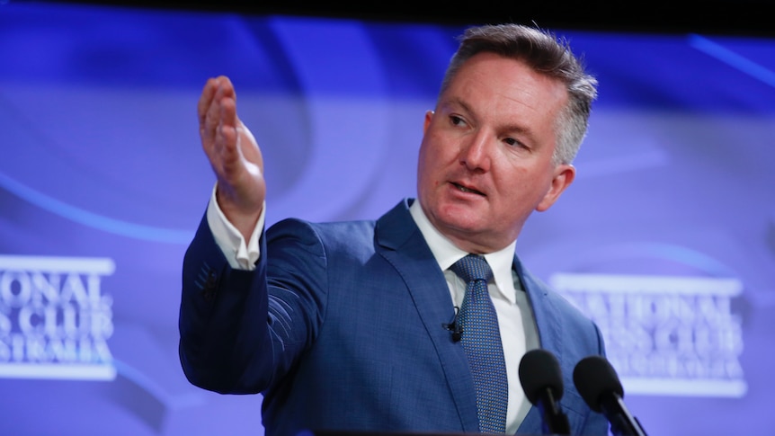 A man wearing a suit gestures as he stands in front of a lectern and a branded backdrop.