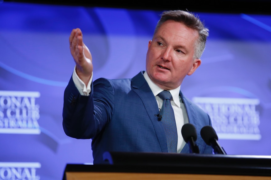 A man wearing a suit gestures as he stands in front of a lectern and a branded backdrop.