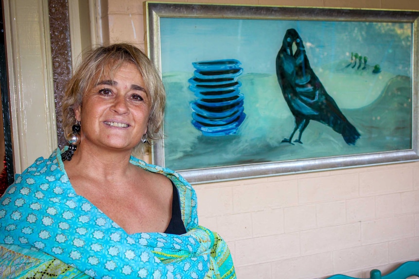 A woman poses beside a painting of a bower bird and a stack of dishes.
