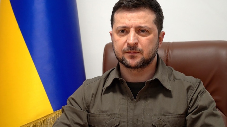 Volodymyr Zelensky sits in a brown leather chair with a Ukrainian flag behind him