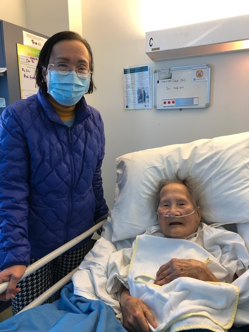 A woman in blue jacketand mask next to older woman in hospital bed.