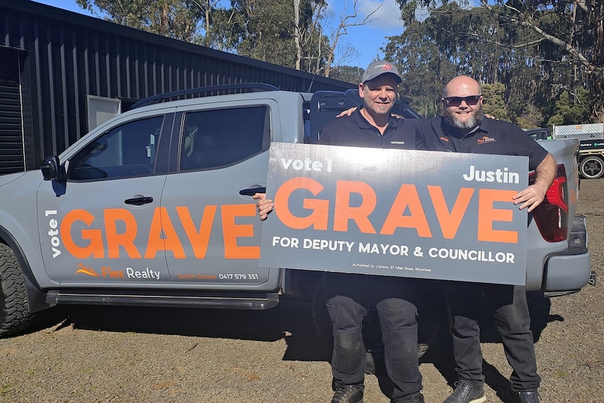 Two young men stand on a sunny day stand in front of a big ute branding with the name "GRAVE" as well as a political sign.