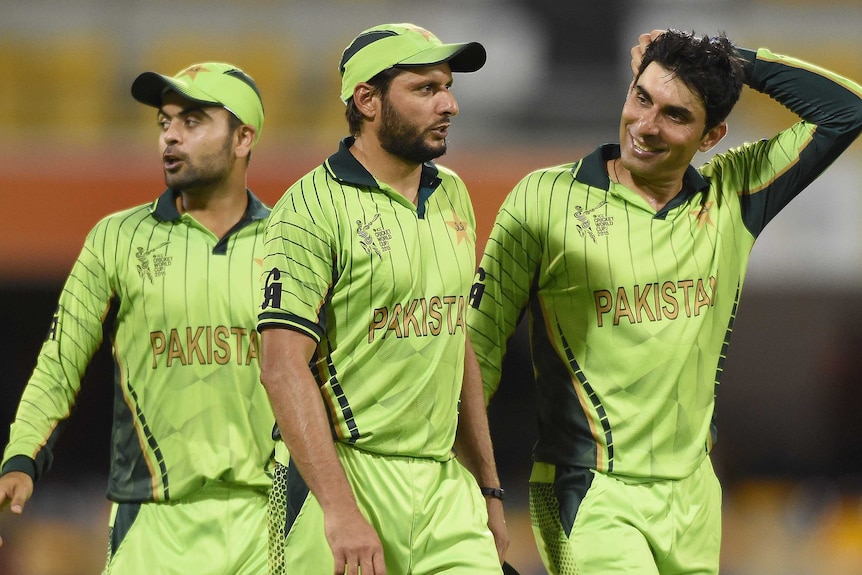 Shahid Afridi stands next to two teammates in lime-green wearing a green cap
