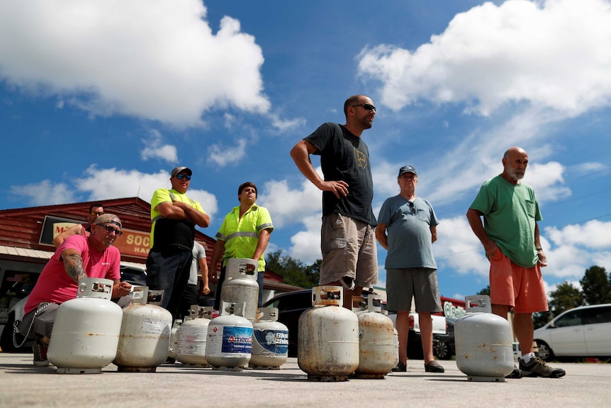 A group of men stand and crouch in a queue with gas canisters at their feet, with blue sky and clouds overhead.