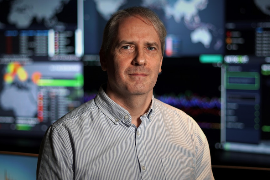 a man in a business shirt with computer screens behind him