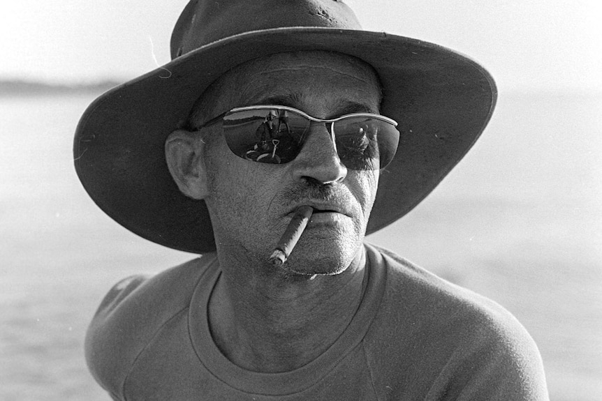 Black and white photo of man wearing a wide-brimmed hat and sunglasses and smoking a cigar