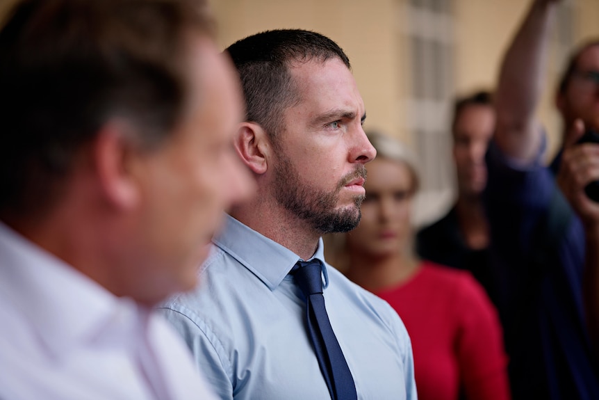 A close-up of caucasian man in a light blue shirt and blue tie, among a crowd of people.