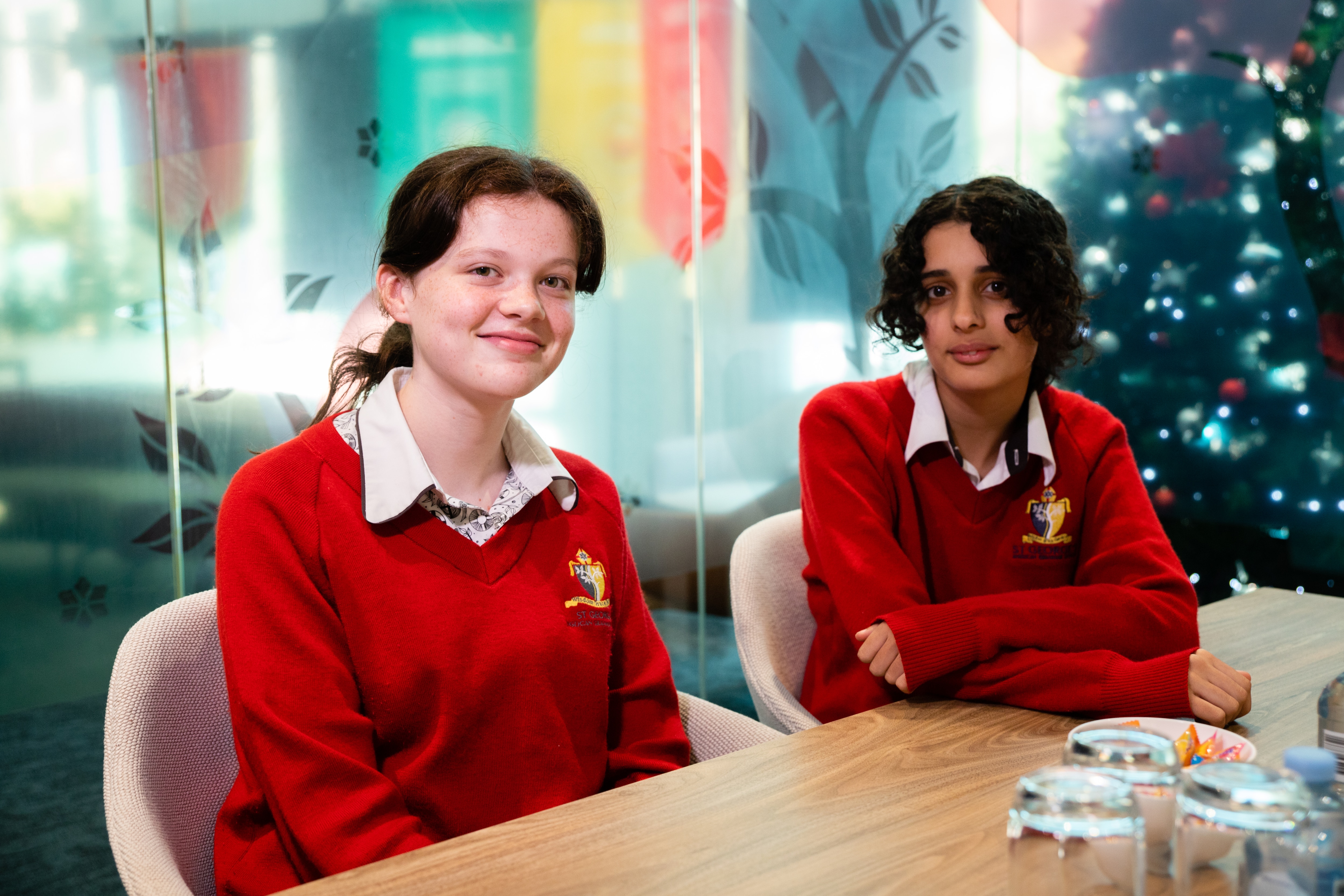 Two young teenage girls sit at a table and smile shyly at the camera in school uniform.