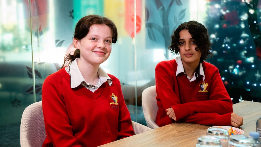 Two young teenage girls sit at a table and smile shyly at the camera in school uniform.