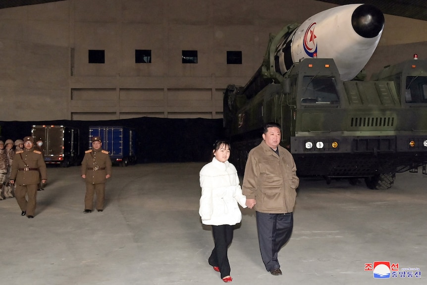 Kim Jong Un marche et tient la main de sa fille, qui porte une chemise blanche, un pantalon blanc et des chaussures rouges.