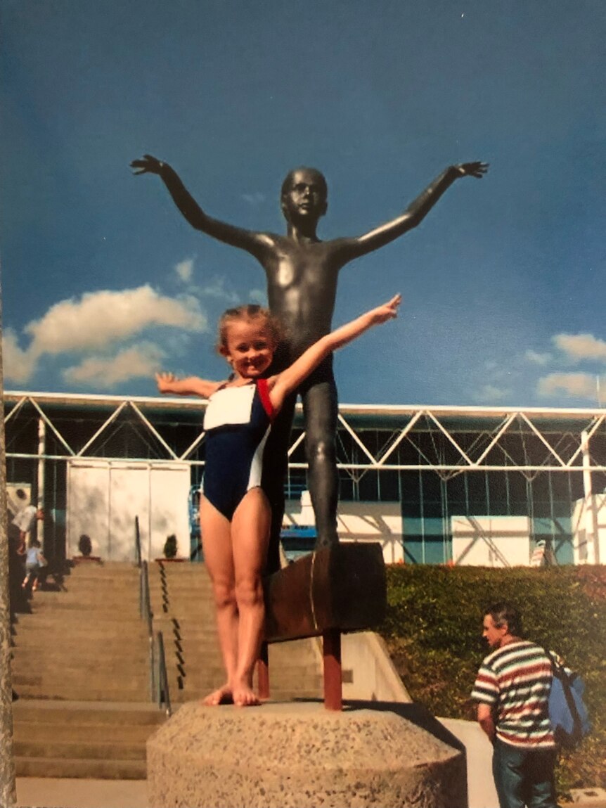 A young girl stands on the podium of a statue, both her and the bronze statue of a young girl behind her with their arms out.