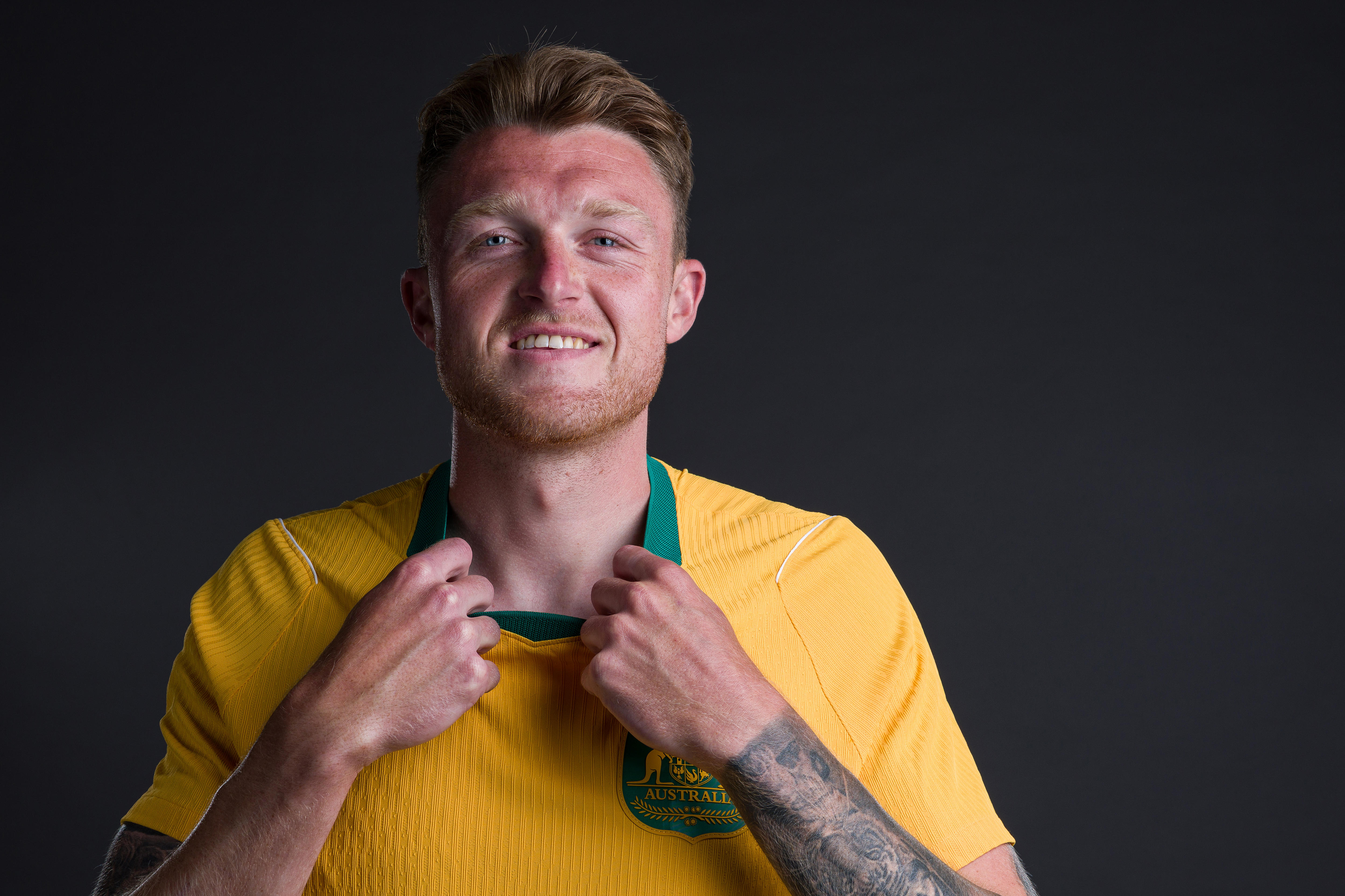 Harry is in yellow and green jersey, holds collar, smiles, has tattoos on his left arm. Background dark.