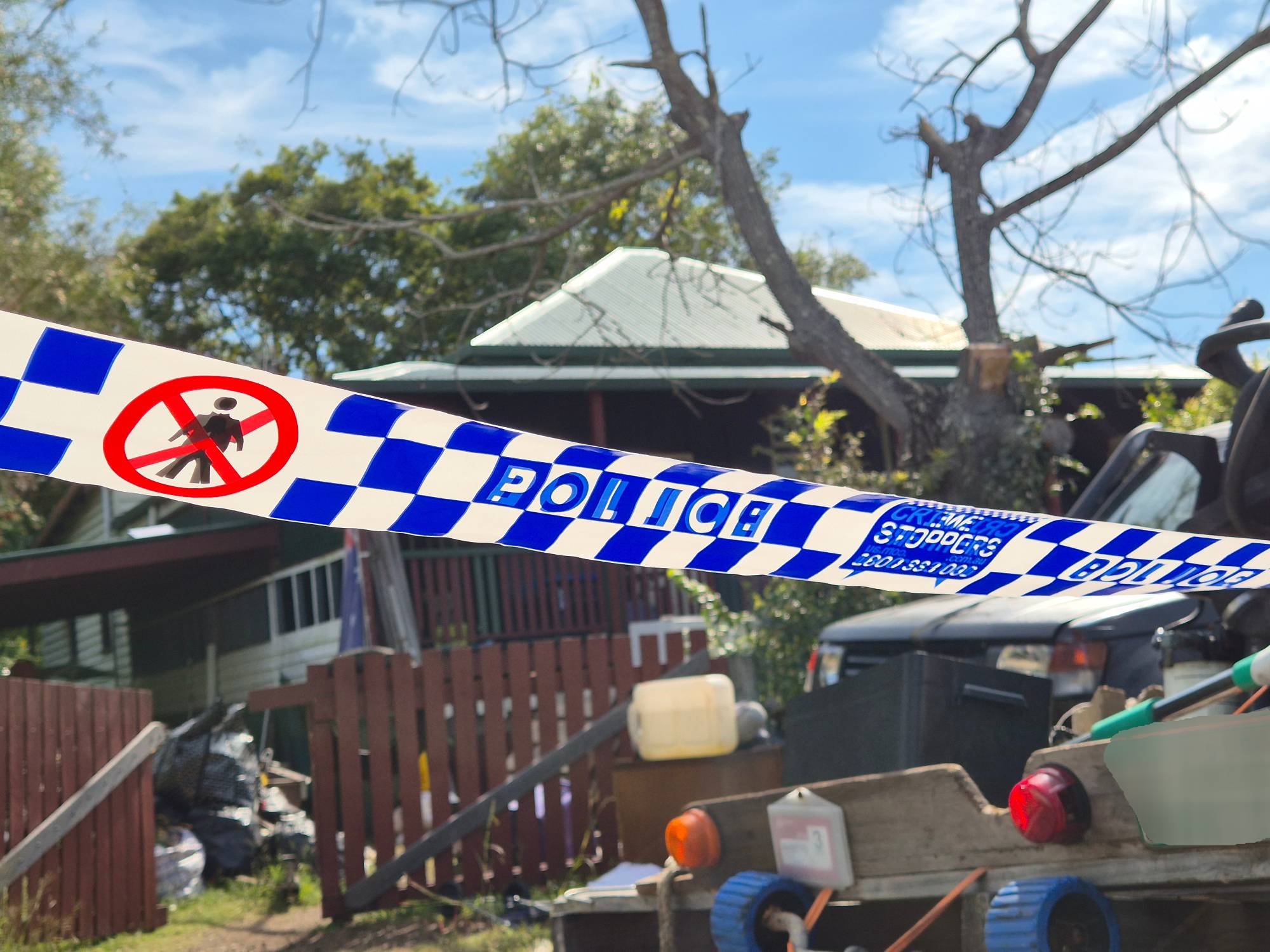 Police barrier tape surrounds a house with a red fence; the yard contains vehicles and scattered debris.