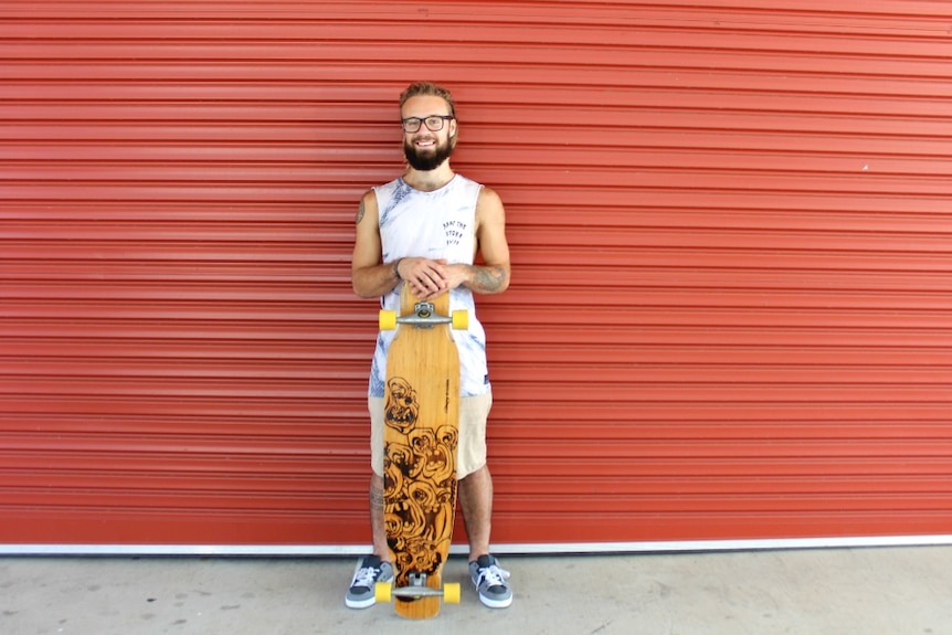 Man with dreadlocks, beard, glasses standing against red roller garage door with a longboard skateboard.
