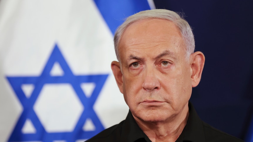 Israel Prime Minister Benjamin Netanyahu looks serious as he stands in front of a blue and white Israeli flag.