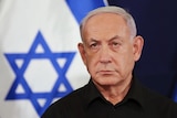 An older man with white hair looks serious as he stands in front of a blue and white Israeli flag.