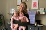 A white woman with long blonde hair stands in an office holding two babies in pink onesies.