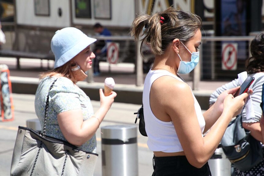At a pedestrian crossing, a woman eats an icecream while another woman in a face mask looks at her phone.
