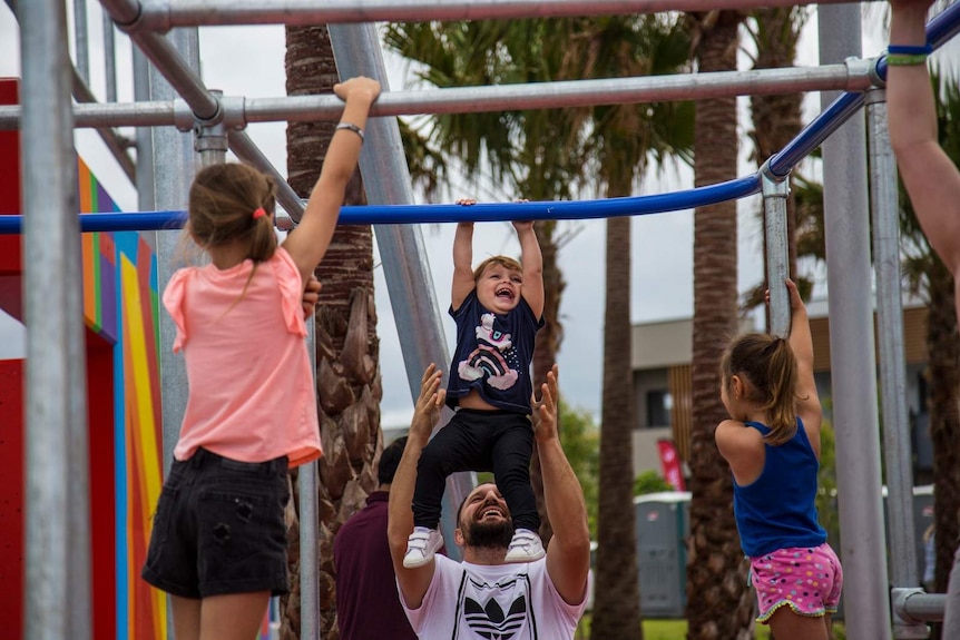 A man holds a young child up to the monkey bars at a park while other children play nearby.