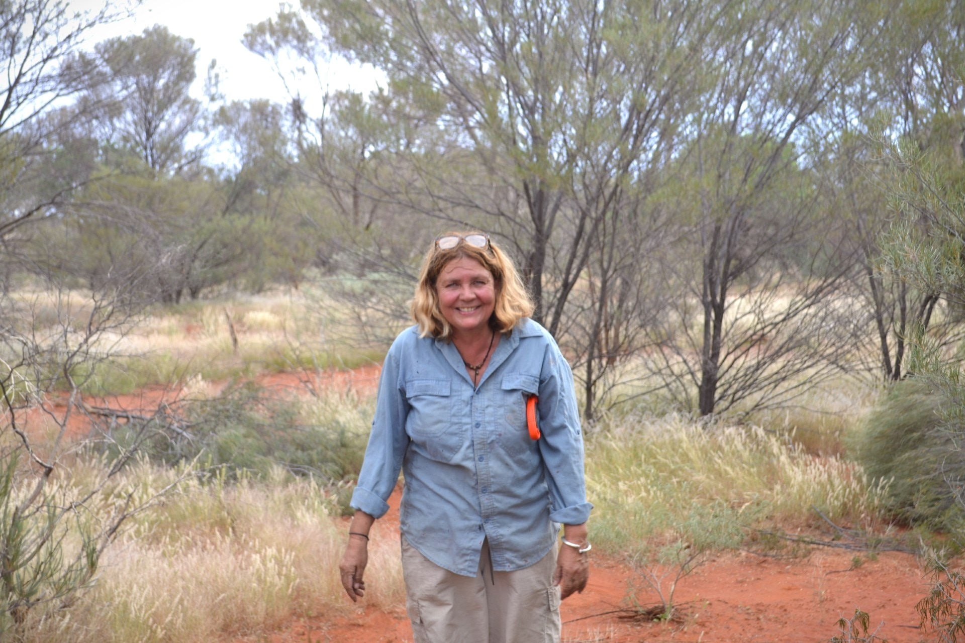 A white woman with shoulder-length blonde hair in blue shirt, walking in Australian bushland.