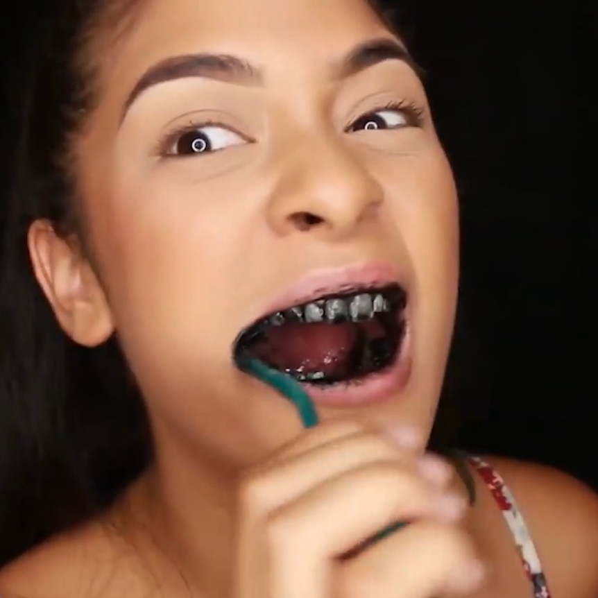 A woman brushes her teeth with charcoal toothpaste, blackening her teeth.