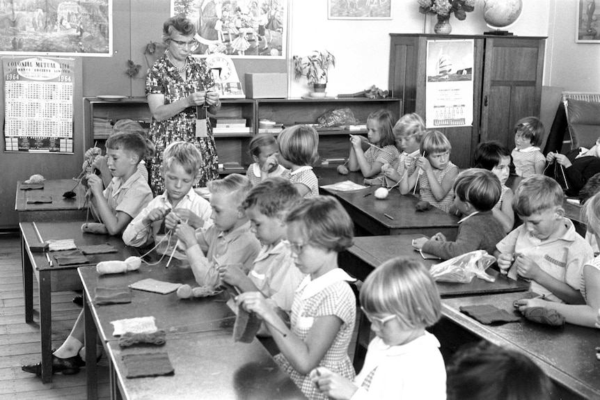 Black and white photo of school children knitting in a classroom with their teacher.