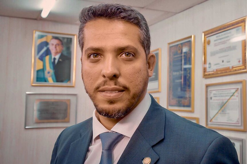 A man in a suit poses for a photograph in an office with a portrait of the Brazilian President in the background