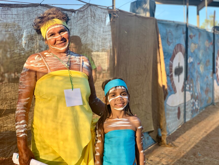 A small girl and a woman stand in the sunlight dressed for Aboriginal junba dance performance