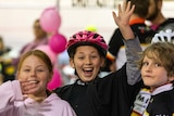 Three children smile and wave, one with a pink bicycle helmet on.