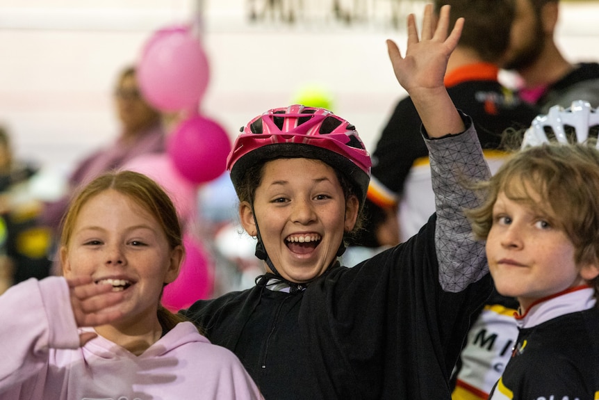 Three children smile and wave, one with a pink bicycle helmet on.