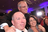 Scott Morrison leans over Garry and Michelle Warren, sitting at a table. They are all smiling at different cameras