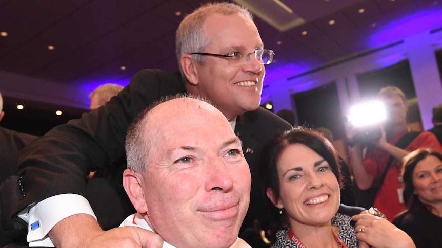 Scott Morrison leans over Garry and Michelle Warren, sitting at a table. They are all smiling at different cameras