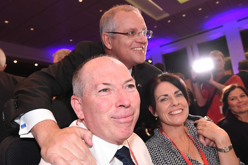 Scott Morrison leans over Garry and Michelle Warren, sitting at a table. They are all smiling at different cameras