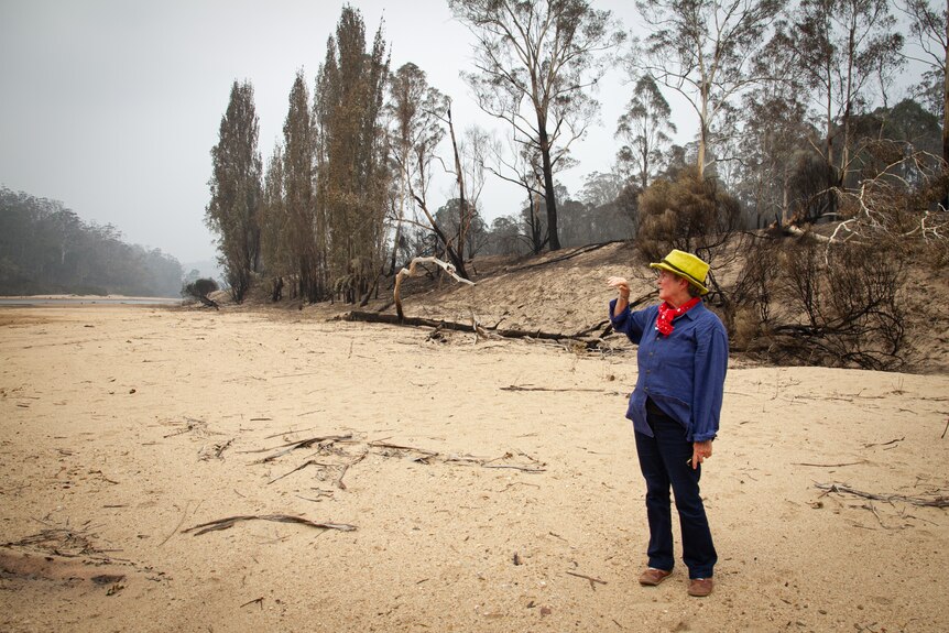 Wide shot of woman standing on a dry riverbed with burnt trees on the bank