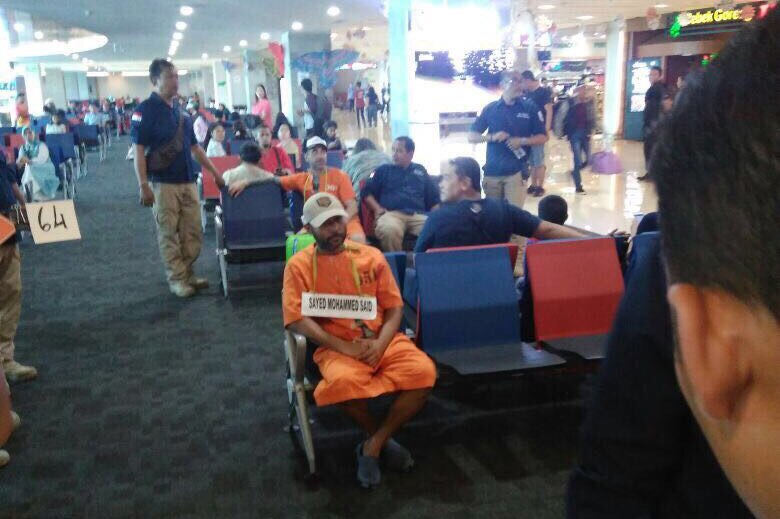 Two prisoners in orange jumpsuits sit in chairs at the Bali airport.