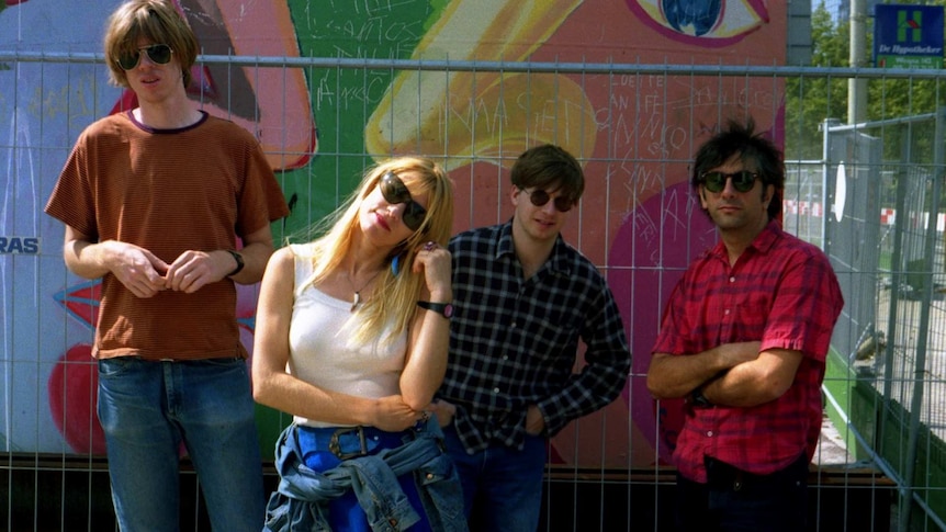 Four members of Sonic Youth stand before a metal fence and colourful mural on an outdoor wall