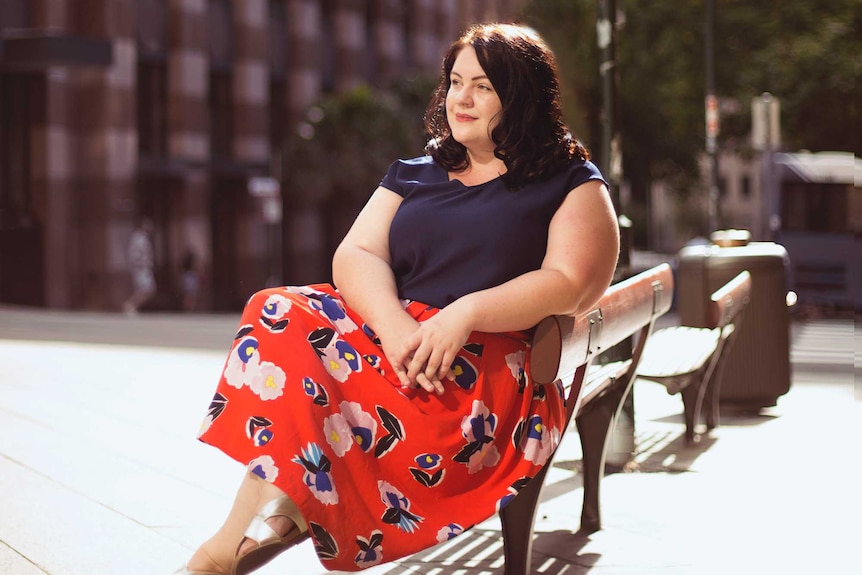 An Aboriginal woman in her 30s wearing a red skirt and navy top sits on a bench in the sun