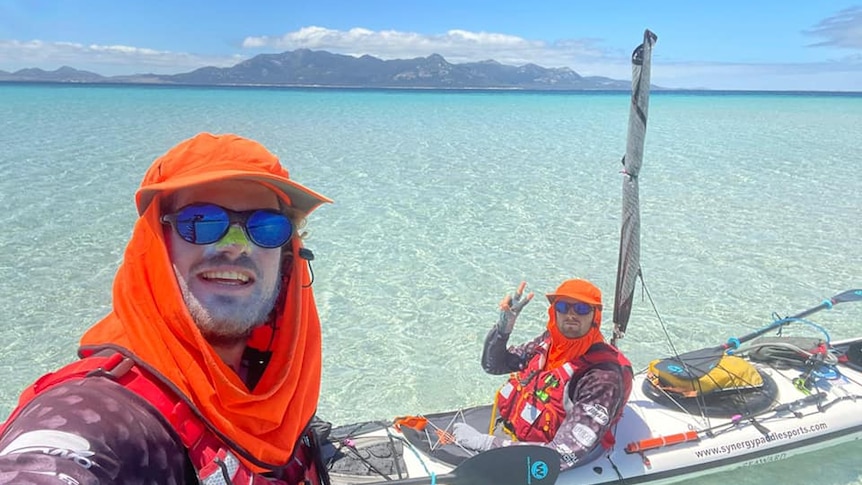two kayakers wearing hats in clear water with an island behind them