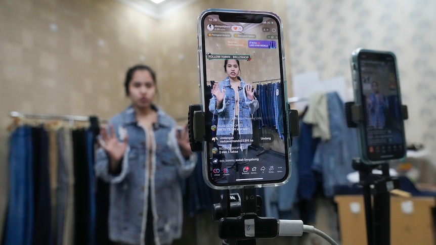 A seller offers merchandise using live streaming over a mouonted mobile phone positioned inside a clothes shop.