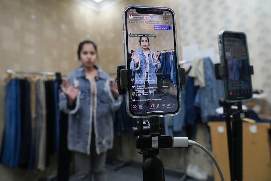 A seller offers merchandise using live streaming over a mouonted mobile phone positioned inside a clothes shop.
