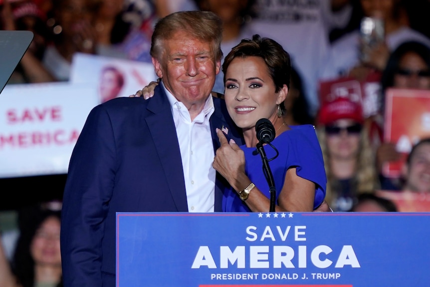 Kari Lake hugs Donald Trump close while pointing at him as he smirks. They stand in front of a SAVE AMERICA lectern