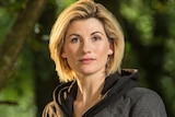 A blonde Jodie Whittaker stands in the woods while removing her hoodie.
