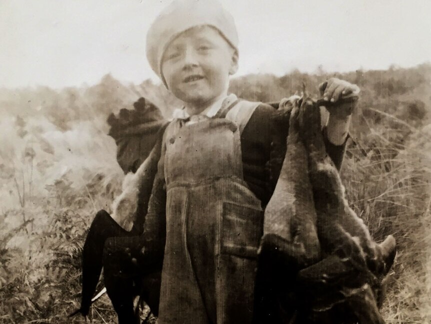 Very old black and white photo of a young boy smiling with a stick of harvested mutton birds across his shoulders.