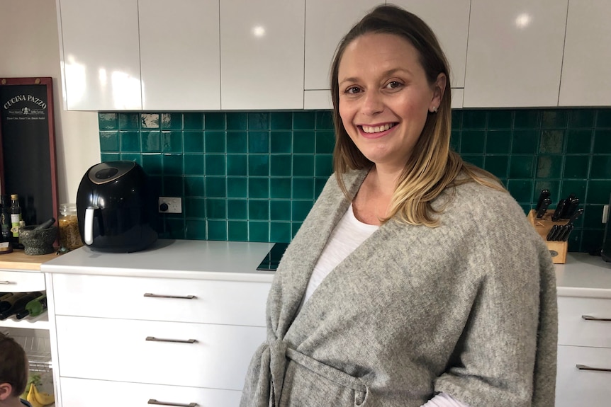 A pregnant woman stands in her kitchen, wearing a grey cardigan.