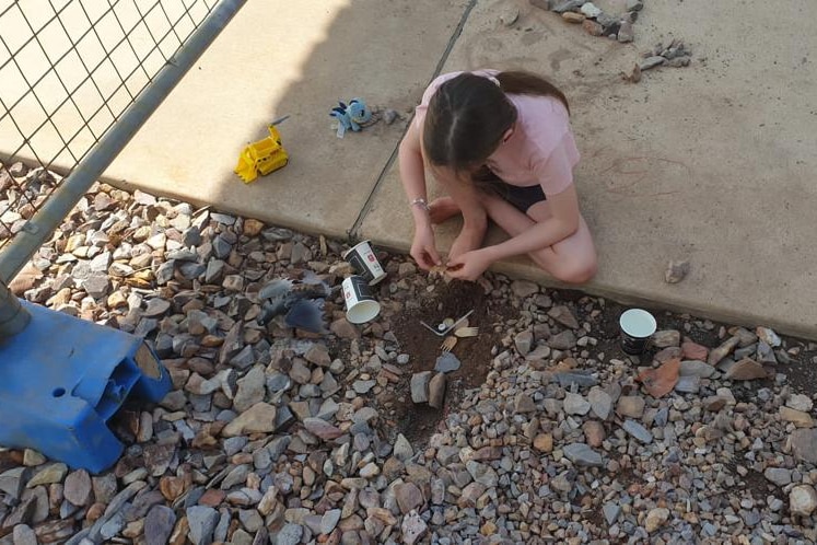 A young girl sits in the dirt playing with some toys and empty coffee cups, next to a steel fence.