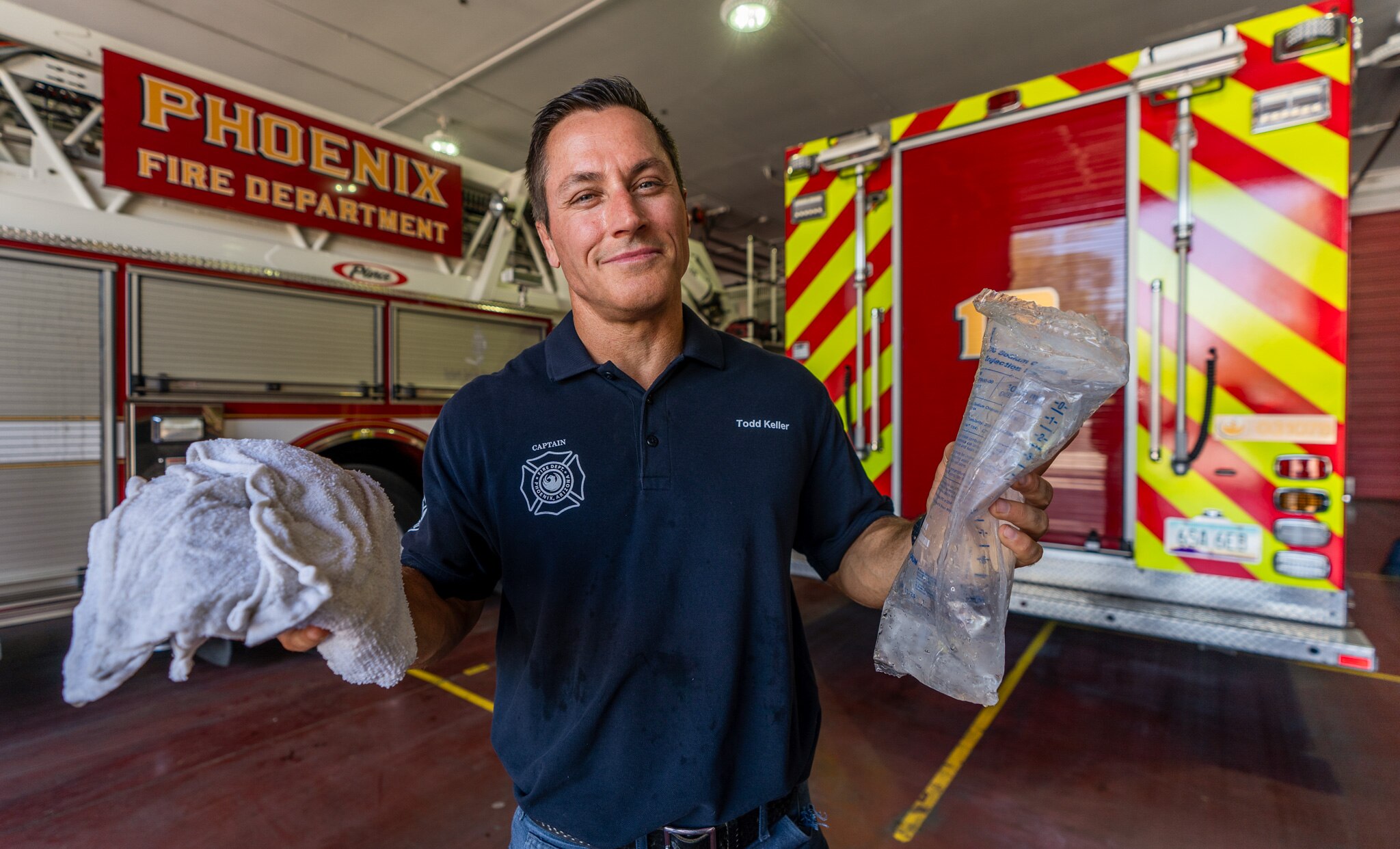 A man wearing a dark blue polo holds up a towel and an IV-fluid bag beside a fire truck.