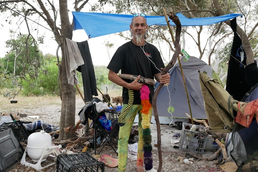 A mostly-bald man with a matted beard, dressed in a black shirt and colourful pants, stands in bush campsite.