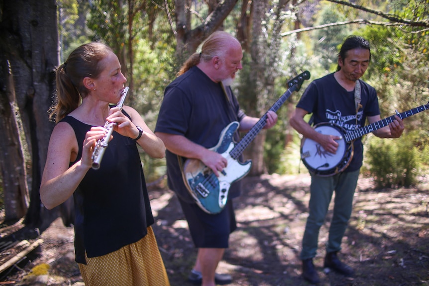 Three people stand playing instruments. Flute, electric guitar and banjo