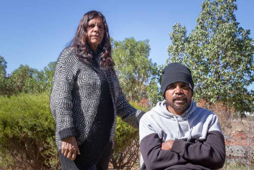 A woman stands behind a seated man outside.